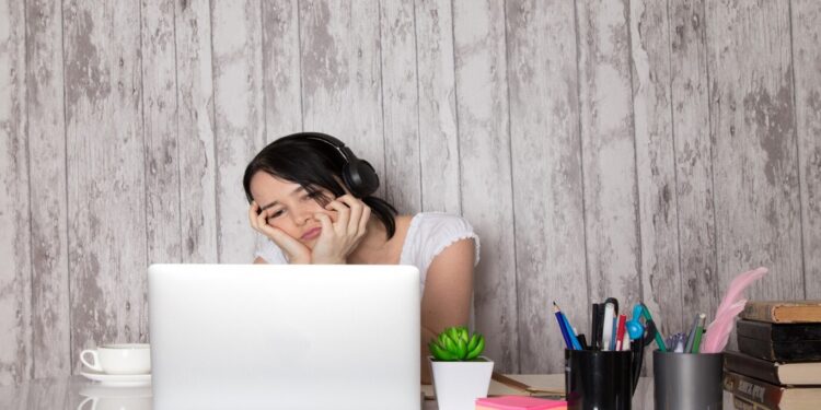 Mulher com fones de ouvido e expressão cansada usando notebook em mesa com itens de escritório