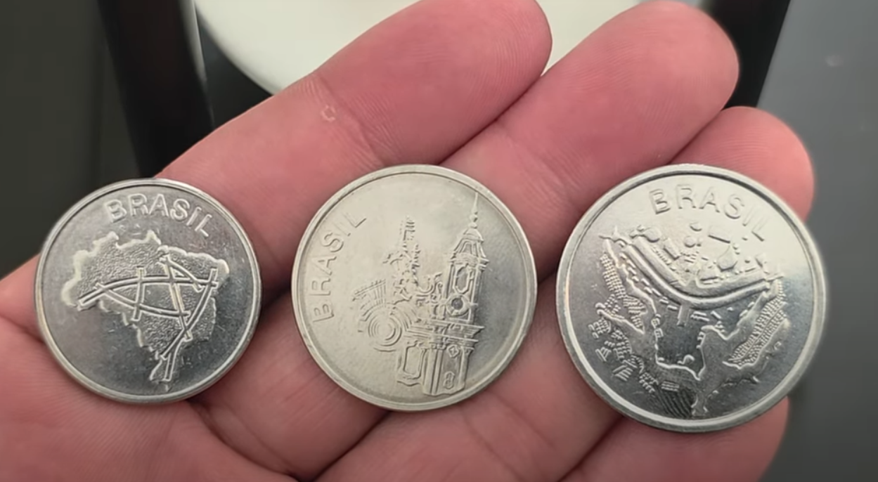 Moedas antigas brasileiras com ilustrações de mapas e monumentos Três moedas brasileiras antigas seguradas na mão, mostrando desenhos e mapas do Brasil