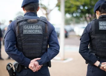Dois agentes da Guarda Municipal observando a área pública com uniformes completos e coletes táticos.