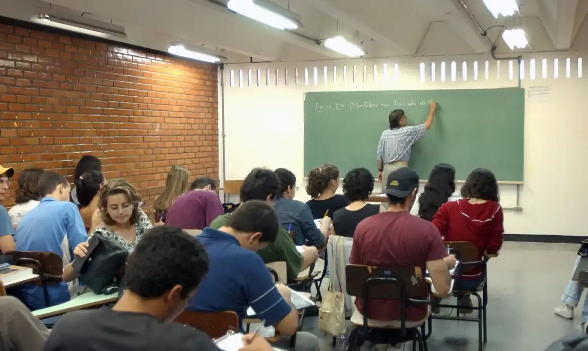 Sala de aula cheia de estudantes atentos enquanto o professor escreve no quadro negro