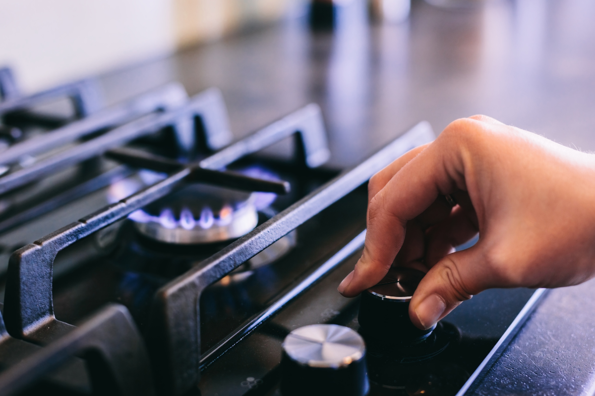 Mão ajustando o botão de um fogão a gás com chama azul acesa.