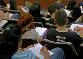 Estudantes concentrados fazendo anotações durante aula em auditório lotado.