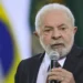 Ex-presidente Lula falando ao microfone durante evento oficial com bandeira do Brasil ao fundo.
