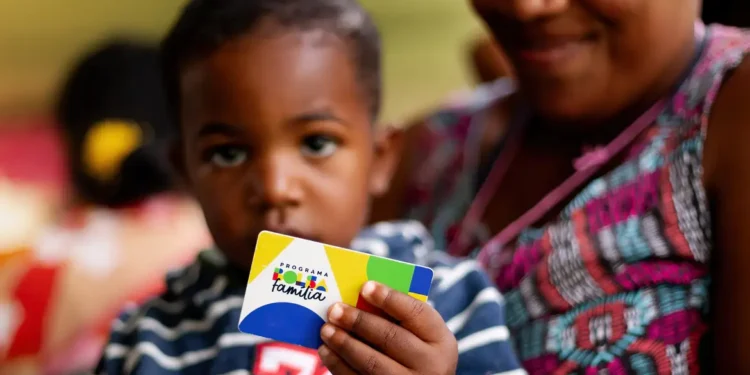 Criança segurando cartão do Programa Bolsa Família, com mãe ao fundo.