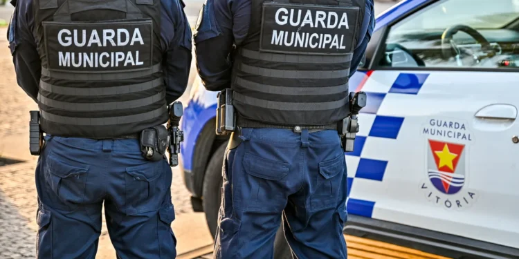 Dois agentes da guarda municipal com coletes táticos vistos de costas durante patrulha urbana.
