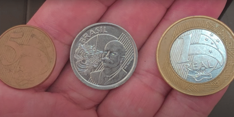 Três moedas brasileiras raras sendo exibidas na palma da mão: 5 centavos, 1 real e comemorativa