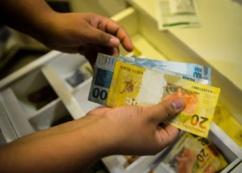 Pessoa segurando cédulas de R$ 100 e R$ 20 em mãos, organizando dinheiro em gaveta.