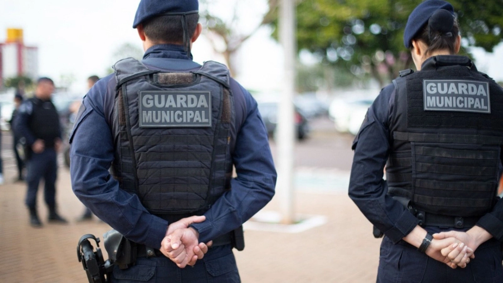 Guardas municipais em uniformes pretos, com as mãos para trás, em uma área pública.