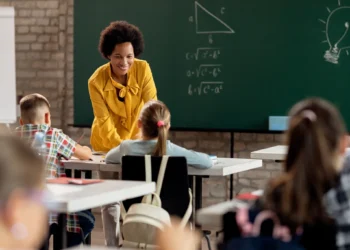 Professora sorrindo e interagindo com alunos em sala de aula, explicando conteúdo em quadro negro.