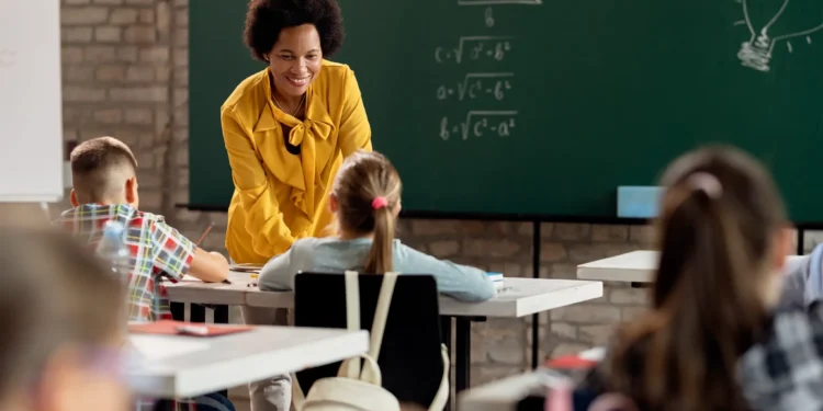 Professora sorrindo e interagindo com alunos em sala de aula, explicando conteúdo em quadro negro.