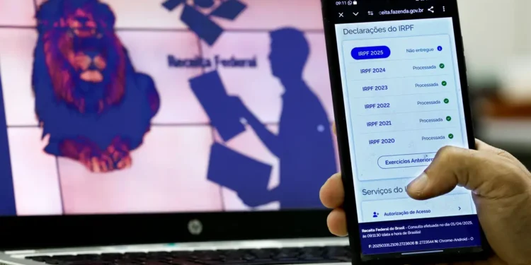 Pessoa segurando celular mostrando consulta de declarações do Imposto de Renda com tela de computador ao fundo exibindo logotipo da Receita Federal
