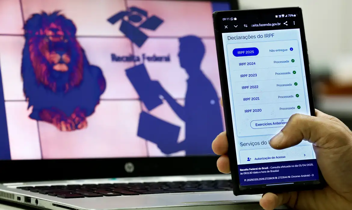 Pessoa segurando celular mostrando consulta de declarações do Imposto de Renda com tela de computador ao fundo exibindo logotipo da Receita Federal
