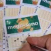 Pessoa marcando números em um volante da Mega-Sena com várias apostas ao fundo.
