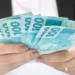 Pessoa segura várias cédulas de 100 reais nas mãos.