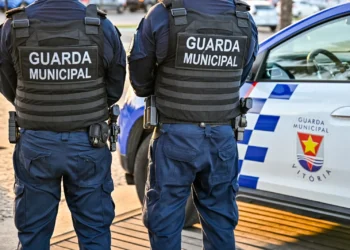Dois agentes da Guarda Municipal de costas ao lado de viatura oficial.