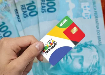 Pessoa segurando cartão do Bolsa Família diante de notas de R$100.