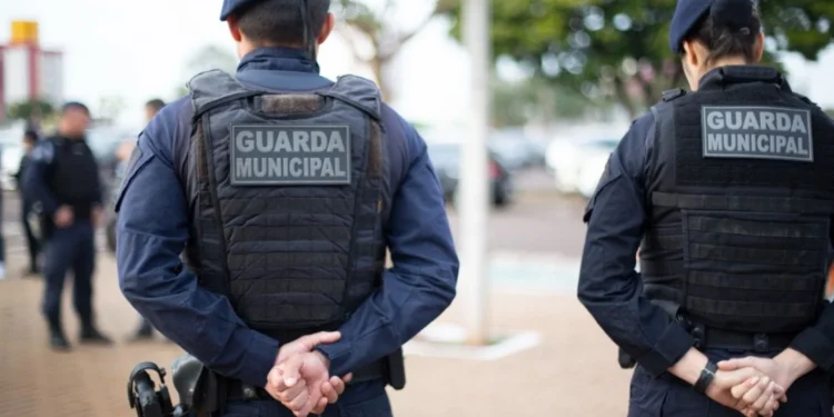 Dois policiais da Guarda Municipal observando a cena, com suas vestes de proteção visíveis e uniformes de segurança.