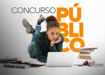 Jovem concentrada estudando online para concurso público, rodeada por livros.