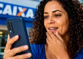 Mulher consulta o celular em frente a uma agência da Caixa, representando a solicitação do Saque Calamidade do FGTS pelo aplicativo.