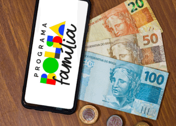 Smartphone exibindo logo do Programa Bolsa Família ao lado de cédulas de 20, 50 e 100 reais com moedas brasileiras