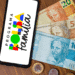 Smartphone exibindo logo do Programa Bolsa Família ao lado de cédulas de 20, 50 e 100 reais com moedas brasileiras
