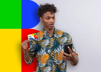 Homem segurando cartão do Bolsa Família e olhando para o celular, com fundo colorido em verde, azul, amarelo e vermelho.