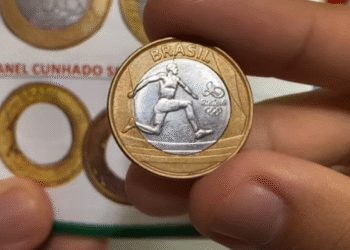 A mão de uma pessoa segurando uma moeda olímpica do atletismo de 1 real, mostrando o salto com vara, com o catálogo de moedas ao fundo.