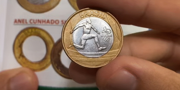 A mão de uma pessoa segurando uma moeda olímpica do atletismo de 1 real, mostrando o salto com vara, com o catálogo de moedas ao fundo.