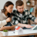 A imagem de um casal analisando plantas de uma casa, com um modelo de miniatura de uma casa na mesa, simbolizando o planejamento da compra de um imóvel.