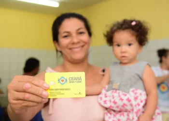 Mulher segurando o cartão do programa Ceará Sem Fome, com uma criança nos braços em um ambiente interno, enquanto outros moradores aparecem ao fundo.