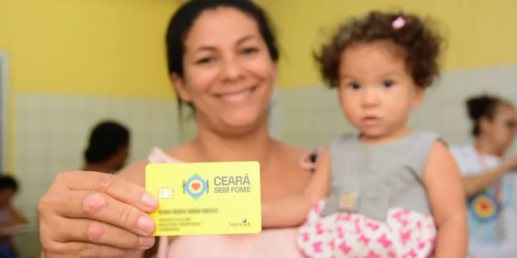 Mulher segurando o cartão do programa Ceará Sem Fome, com uma criança nos braços em um ambiente interno, enquanto outros moradores aparecem ao fundo.