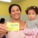 Mulher segurando o cartão do programa Ceará Sem Fome, com uma criança nos braços em um ambiente interno, enquanto outros moradores aparecem ao fundo.