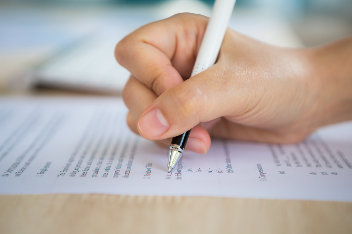 Concursos RJ Mão segurando caneta enquanto assina um documento importante em papel.