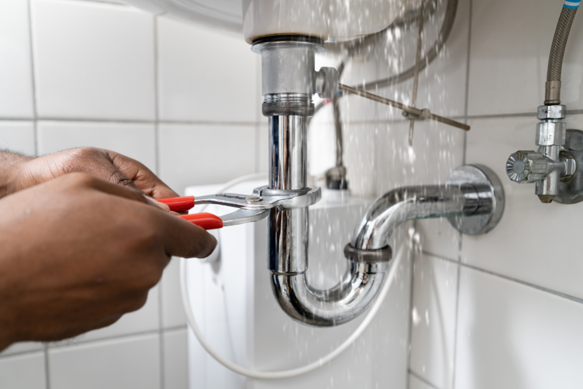 Mãos ajustando cano de pia com vazamento usando alicate, água jorrando.