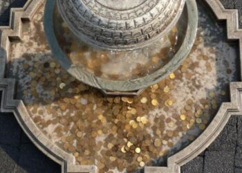 Moedas de cor amarela e prateada acumuladas no fundo de uma fonte de pedra, com a água escorrendo.