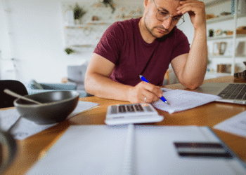 Homem concentrado faz contas financeiras com calculadora, papéis e notebook na mesa.