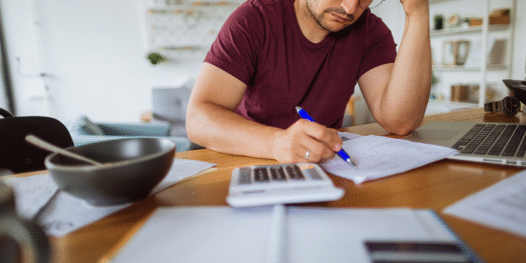 Homem concentrado faz contas financeiras com calculadora, papéis e notebook na mesa.