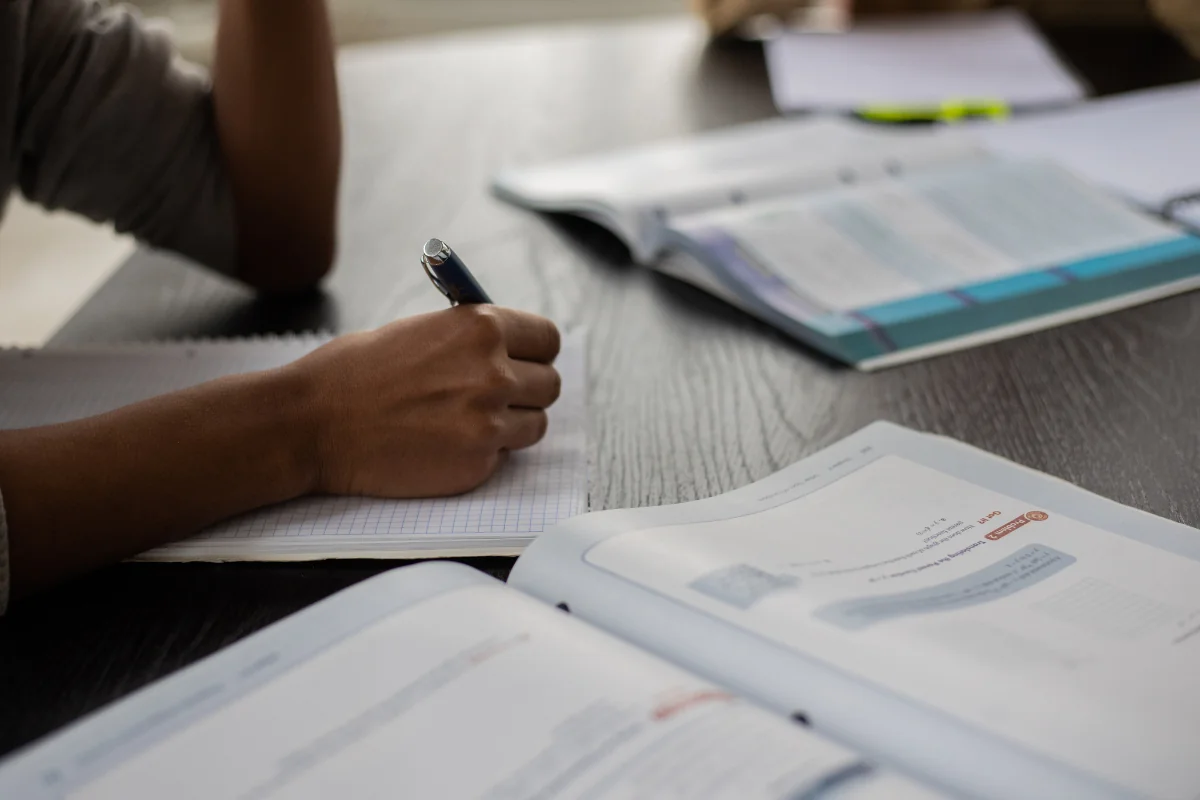 Foco nos estudos com material de apoio aberto durante a preparação Estudante escrevendo em caderno com livros abertos sobre a mesa.
