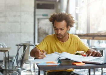 Estudante concentrado estudando com livro aberto e sanduíche ao lado.