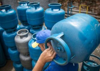 Homem carregando botijão de gás azul em meio a vários botijões, simbolizando a entrega para famílias.
