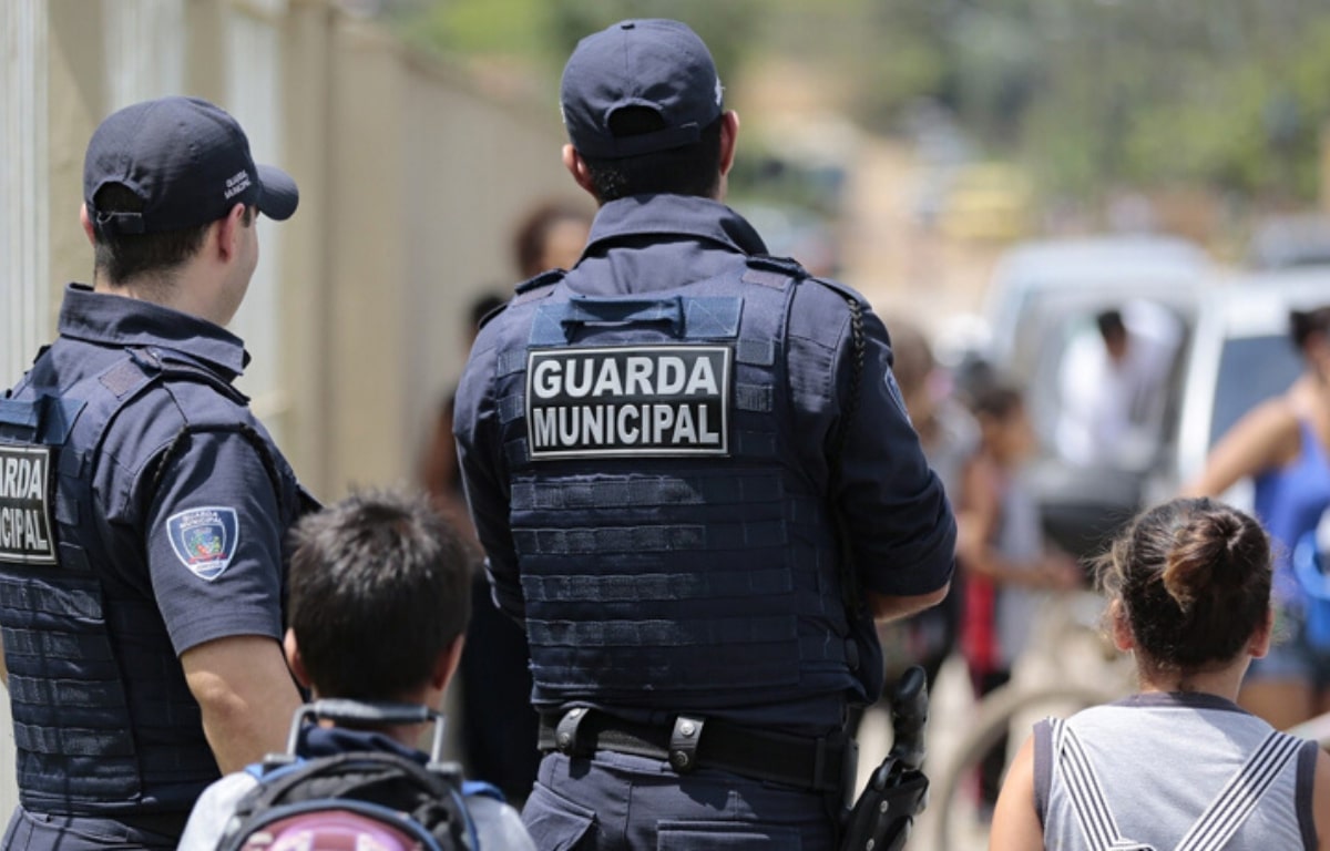 Dois agentes da Guarda Municipal de uniforme observam pessoas caminhando na rua.