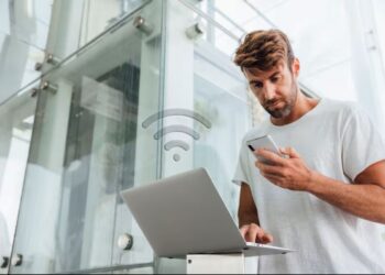 Homem verificando celular e notebook para identificar acesso indevido ao Wi-Fi