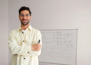 Homem sorrindo, com óculos e camisa clara, em frente a um quadro branco com equações matemáticas e fórmulas, simbolizando o tema de Transferência de curso e reintegração ao Pé-de-Meia Licenciaturas.