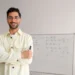 Homem sorrindo, com óculos e camisa clara, em frente a um quadro branco com equações matemáticas e fórmulas, simbolizando o tema de Transferência de curso e reintegração ao Pé-de-Meia Licenciaturas.