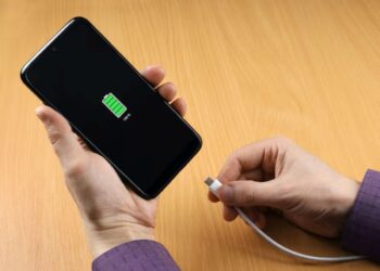 Mãos segurando um celular sendo carregado, indicando bateria cheia.