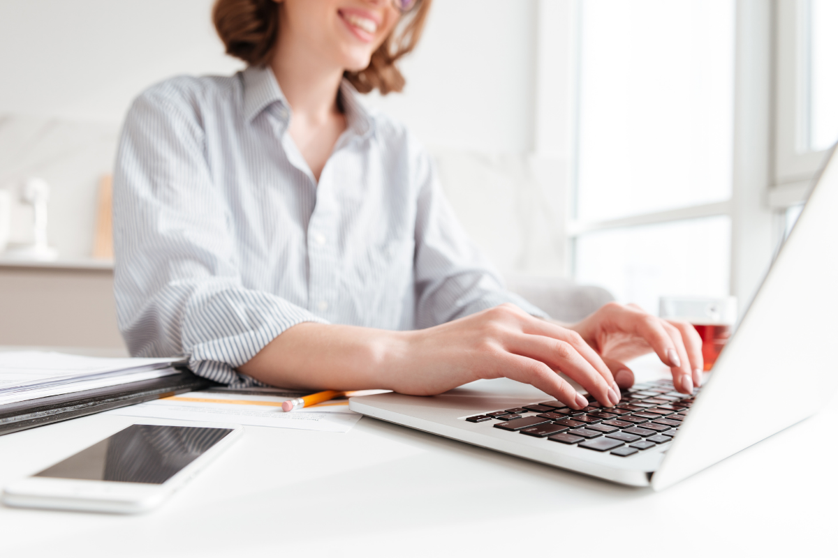 Concurso Prefeitura Mulher digitando em notebook no escritório com smartphone, papéis e lápis na mesa.