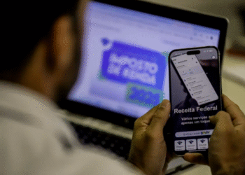 IR 2025: Consulta ao 4º lote de restituição com 3,37% de correção, celular com app Receita Federal e laptop ao fundo.