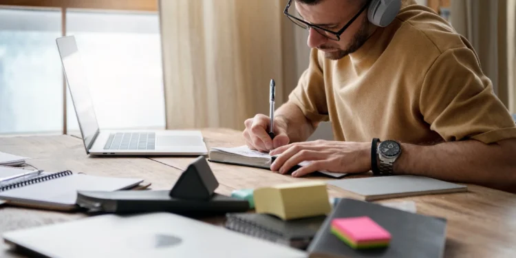 Homem escrevendo em caderno durante sessão de estudo com laptop na mesa.
