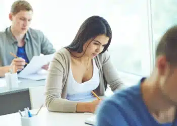 Candidatos concentrados realizando prova de concurso público verificando requisitos do edital