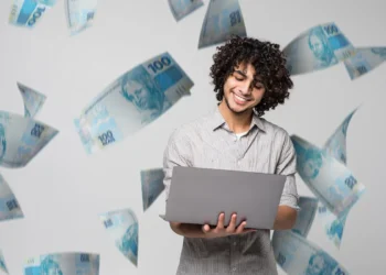 Homem sorrindo com laptop, enquanto notas de dinheiro caem ao seu redor, simbolizando organização financeira.